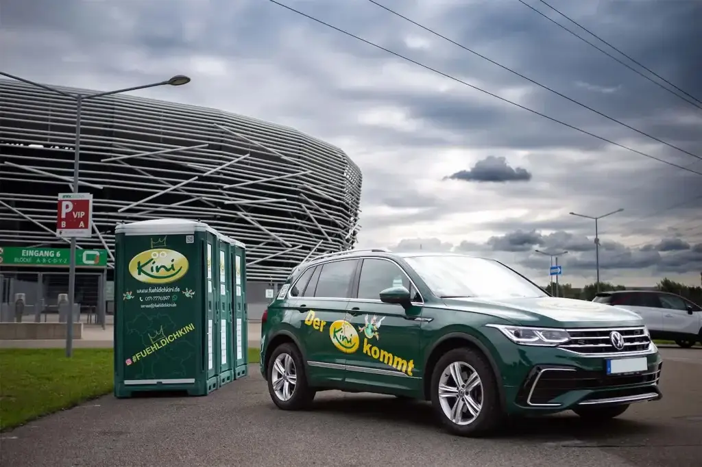 Kini-Auf-Reisen-drei-scheene-mobile-toiletten-vor-sportarena-neben-grünen-volkswagen-tiguan-mit-fuehldichkini-logo