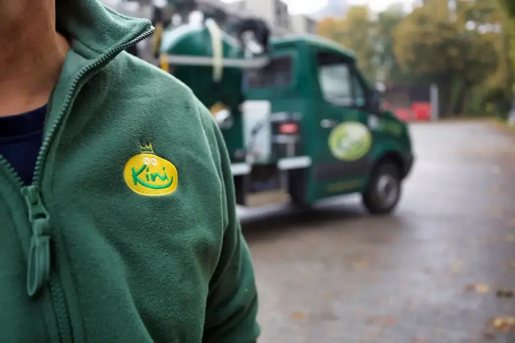 fuehldichkini-kini-heisl-scheenes-dixi-toiletten-service-mitarbeiter-makro-aufnahme-des-sweaters-mit-kini-logo-vor-service-fahrzeug-von-fuehldichkini-in-grün-toilettenwagen-mieten-toiletten-container